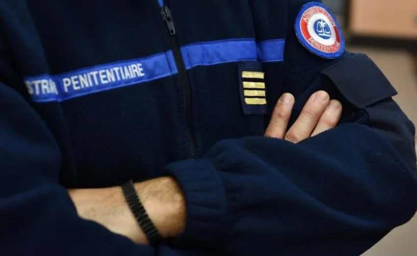 Détail d'un uniforme bleu foncé de l'administration pénitentiaire française, avec un galon bleu et le texte 'STRA PENITENTIAIRE'. Des insignes dorés sont visibles sur le devant.