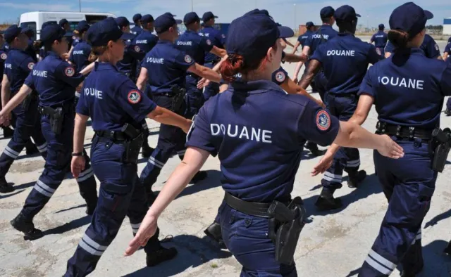 Groupe d'agents de douane en uniforme bleu foncé, certains portant un pistolet à la ceinture, en formation à l'extérieur.
