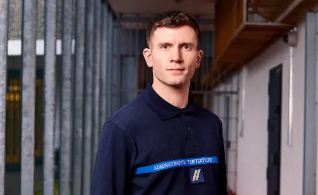 Un homme portant un polo bleu marine de l'administration pénitentiaire, devant des barreaux de prison.