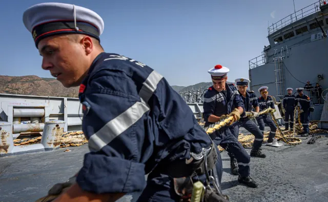Groupe de marins en uniforme bleu tirant ensemble sur une grosse corde sur le pont d'un navire par temps ensoleillé.