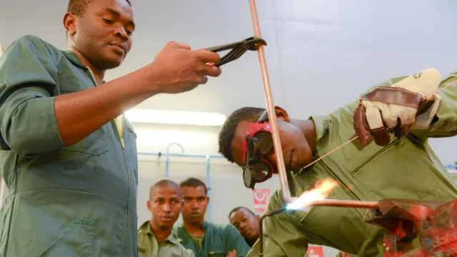 Un apprenti soude un tuyau en cuivre sous le regard d'un instructeur et d'autres étudiants en formation.